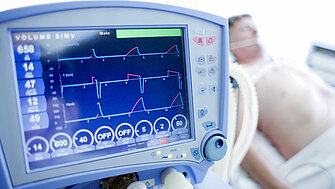 Bildschirm eines Überwachungsmonitors, im Hintergrund liegt ein Patient im Krankenhausbett
