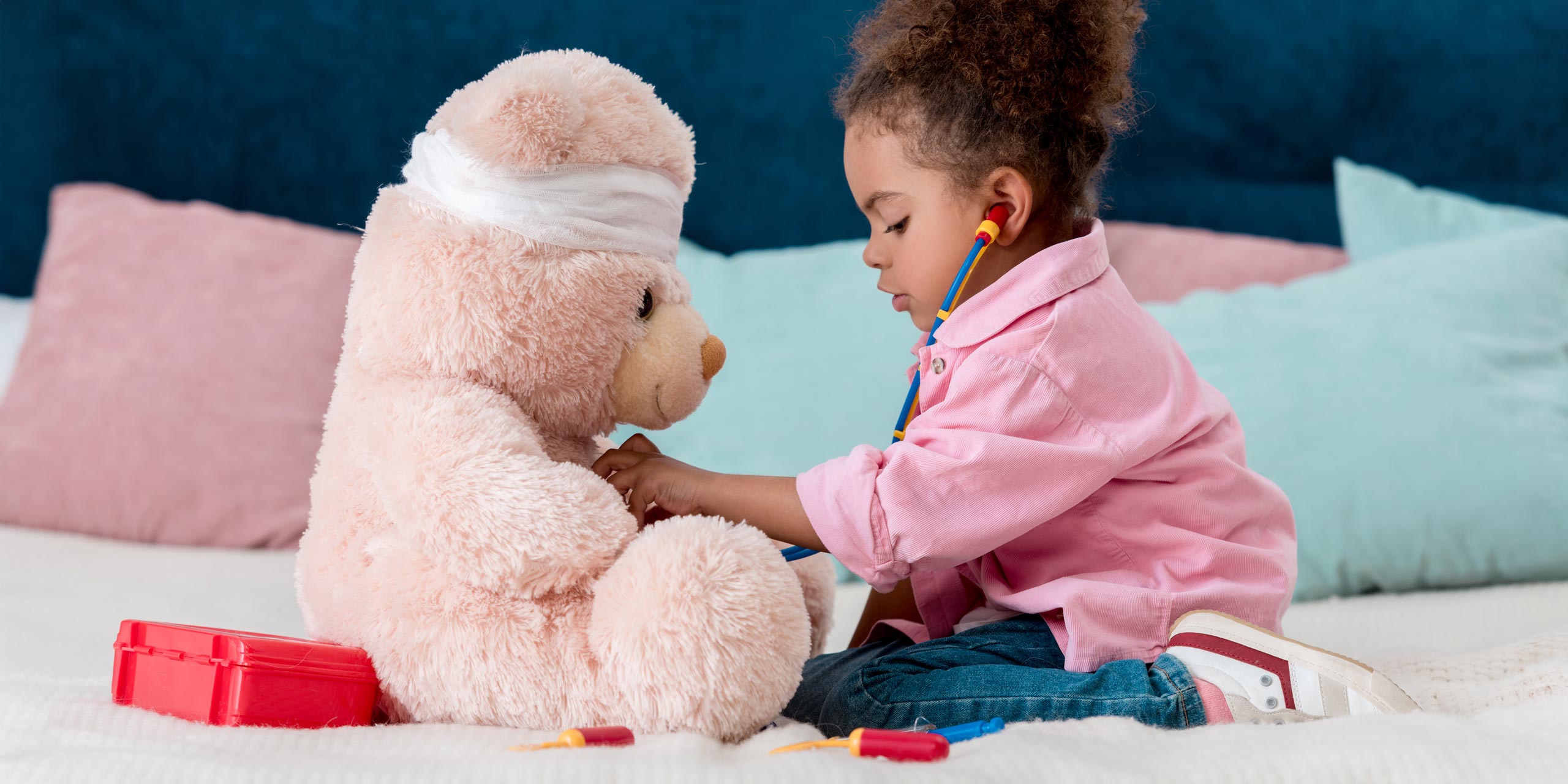 Kleines Mädchen mit Spielzeug-Stethoskop beim Ärztinspielen für Ihren großen rosafarbenen Plüschbären