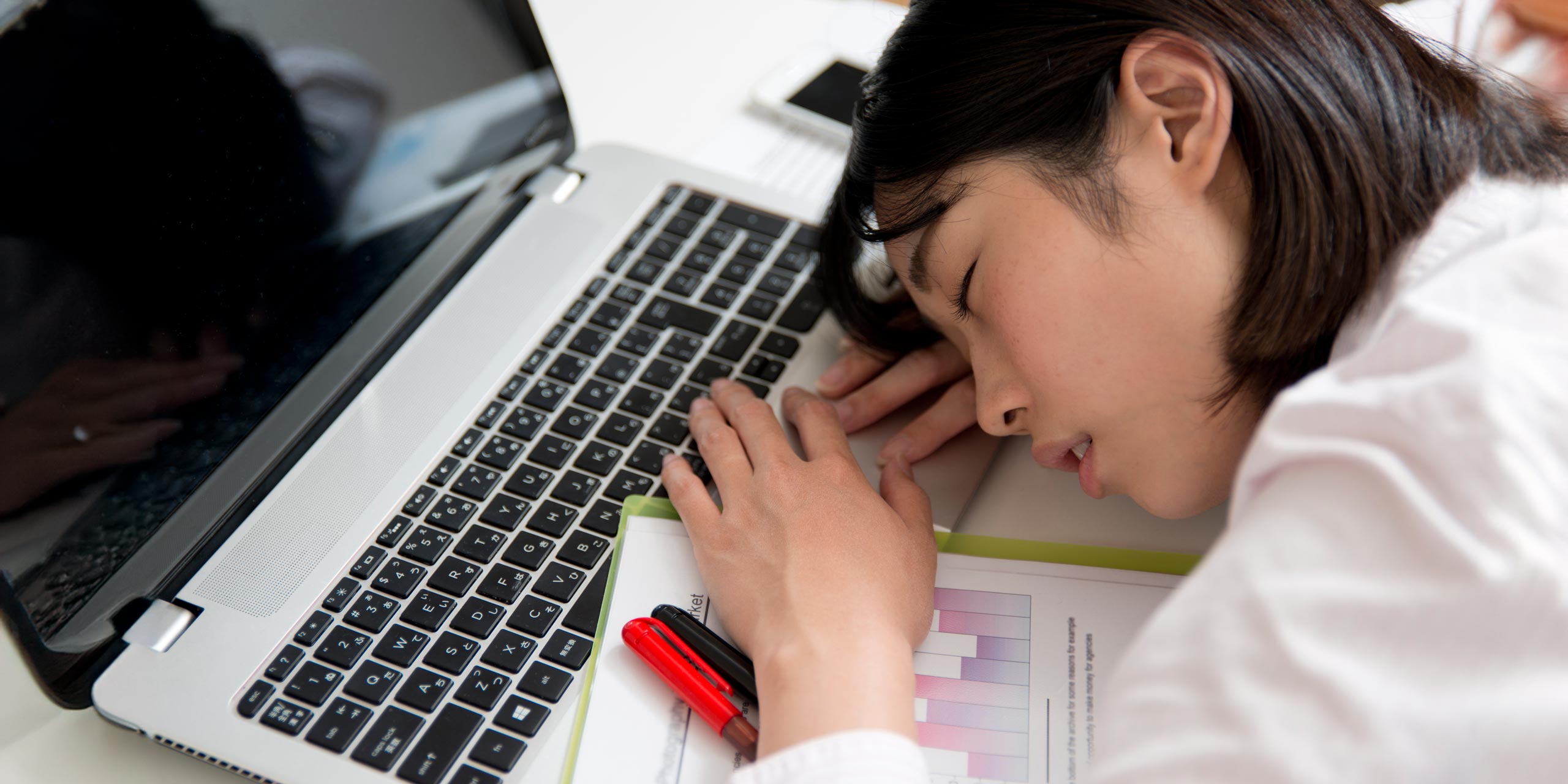 Eine junge Asiatin in Büro-Bekleidung, schlafend, vor ihrem Notebook.